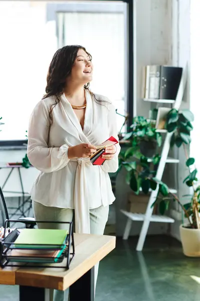 A confident young woman smiles at office, holding a book and surrounded by plants. — Stock Photo