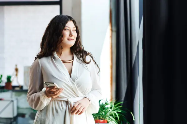 A beautiful plus size woman stands in her cozy office, smiling softly as she looks out the window. — Stock Photo