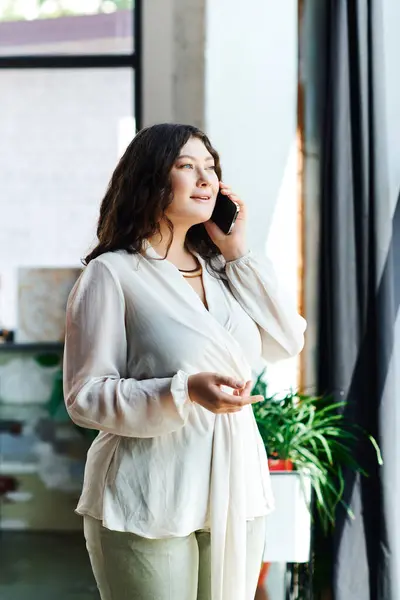 A young, beautiful plus size woman stands by a large window, enjoying a joyful chat. — Stock Photo