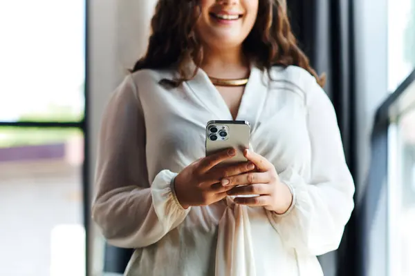 A plus size woman in a cozy atmosphere smiles as she engages on her smartphone at office. — Stock Photo