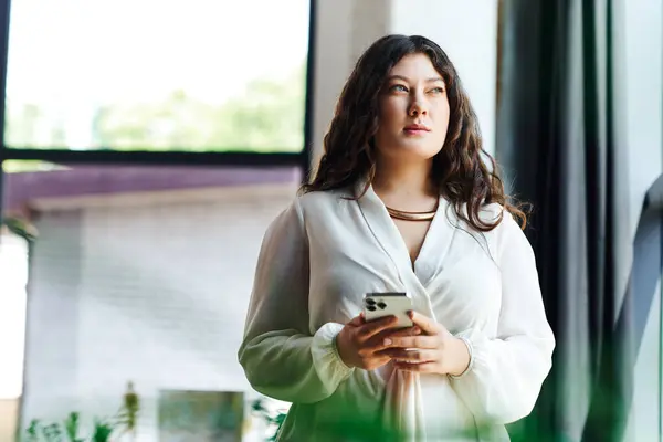 A young plus size woman stands thoughtfully at office, holding her phone and gazing outside. — Stock Photo