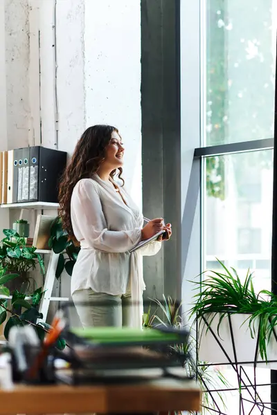 A young beautiful plus size woman stands by the window, enjoying a serene moment in her office. — Stock Photo