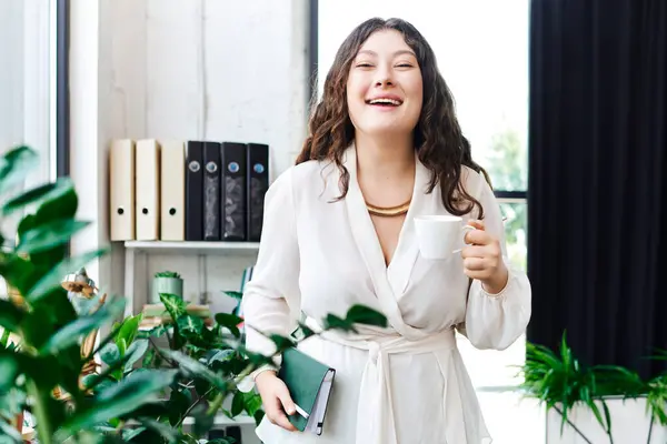 A plus size woman smiles brightly while holding a cup of coffee in a cozy office environment. — Stock Photo