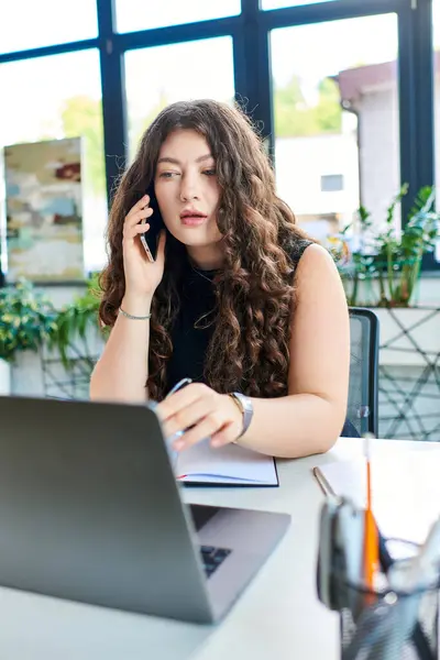 A young plus size professional with long curly hair focuses intently while working on her laptop in a vibrant office environment. — Stock Photo