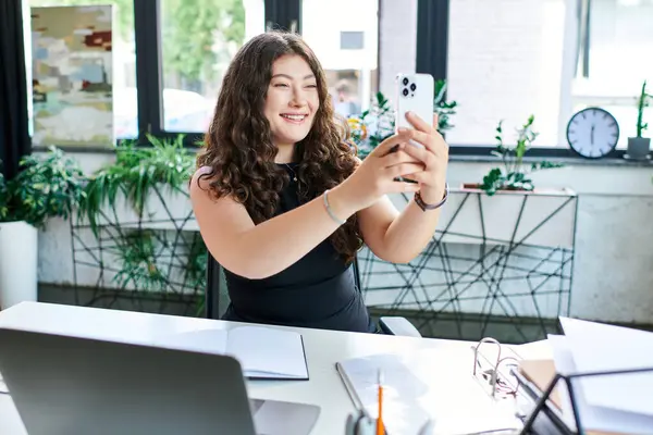 Una alegre mujer de negocios de tamaño grande sonríe mientras toma una videollamada en su elegante oficina rodeada de plantas. - foto de stock
