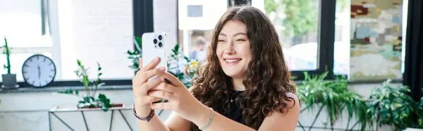 Ein Plus-Size-Profi mit langen lockigen Haaren lächelt, während er ein Selfie in einem lebhaften Bürobereich mit viel Grün macht. — Stockfoto