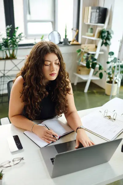 Una donna fiduciosa plus-size in un ufficio elegante scrive note mentre si impegna con il suo computer portatile, incarnando la cultura aziendale. — Foto stock