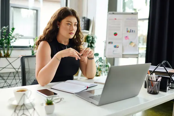 A confident plus-size woman with long curly hair focuses on her laptop in a bright office, surrounded by plants. — Stock Photo