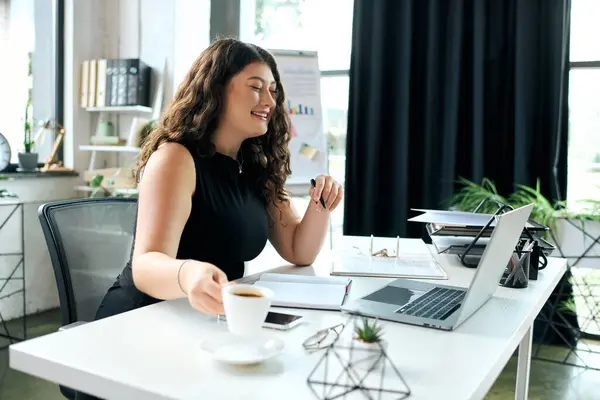 A young plus-size woman with curly hair smiles as she sips coffee at her desk, immersed in her work in a bright office. — Stock Photo