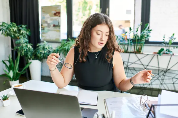 A confident plus-size woman with long curly hair is actively working at her desk, immersed in her tasks. — Stock Photo
