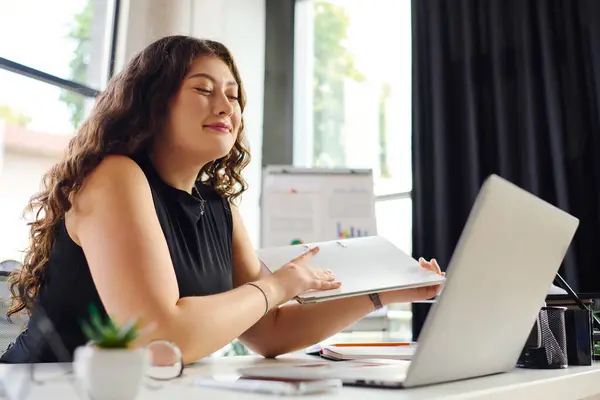 In a bright office, a plus-size woman with long curly hair smiles contentedly while working on her laptop. — Stock Photo