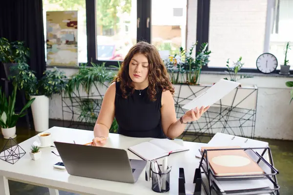 A confident young businesswoman focuses intently on her laptop while reviewing documents in a stylish office. — Stock Photo
