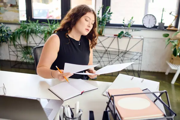 Уверенная в себе женщина plus size работает с документами за стильным офисным столом, демонстрируя свой профессиональный блеск и креативность. — стоковое фото