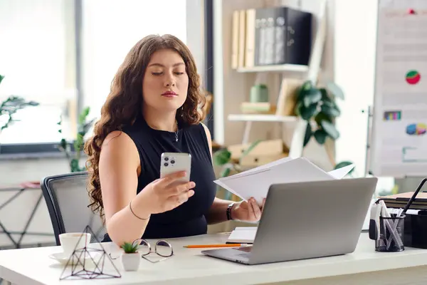 Una joven mujer de talla grande con cabello largo y rizado revisa documentos mientras revisa su teléfono en un elegante escritorio de oficina. — Stock Photo