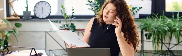 A confident young woman with long curls multitasks in her office, reviewing documents while speaking on her phone, immersed in her work. — Stock Photo