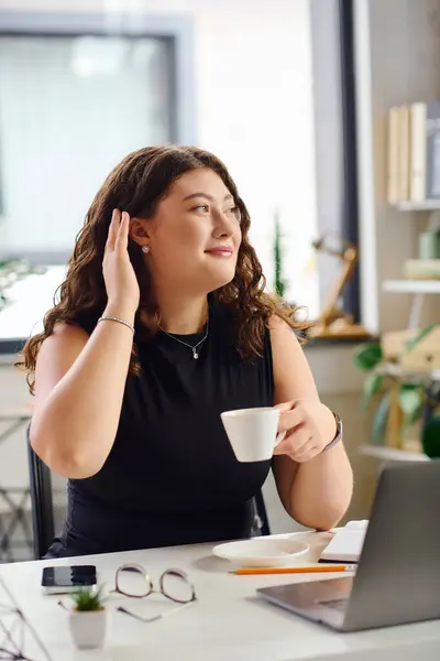 A confident plus size woman with curly hair savors a coffee while taking a moment in her dynamic office environment. — Stock Photo