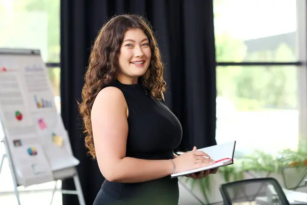 A confident plus size woman with long curly hair stands in a vibrant office, taking notes for work. — Stock Photo