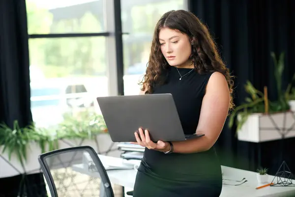 A plus-size woman with long curly hair focuses intently on her laptop in a stylish office setting, showcascasing professionalism. — стоковое фото