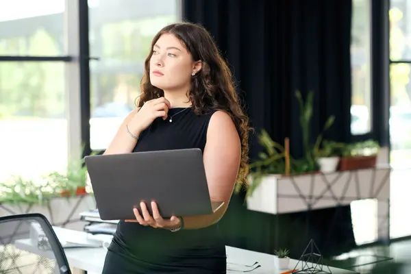 A confident young plus size woman with curly hair is engaged in her work at a modern office, showcasing professional elegance. — Stock Photo