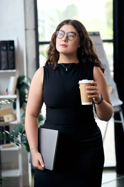 A stylish plus-size woman with curly hair stands confidently in the office, holding her laptop and coffee. — Stock Photo