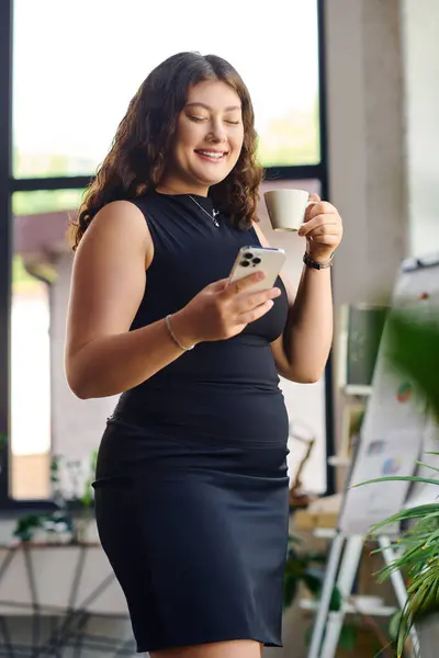 A confident young plus-size woman with curly hair is engaging with her phone while sipping coffee in a vibrant office environment. — Stock Photo