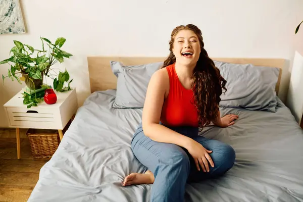 A young woman with curly hair enjoys a moment of happiness while sitting on her bed at home, surrounded by greenery. — Stock Photo