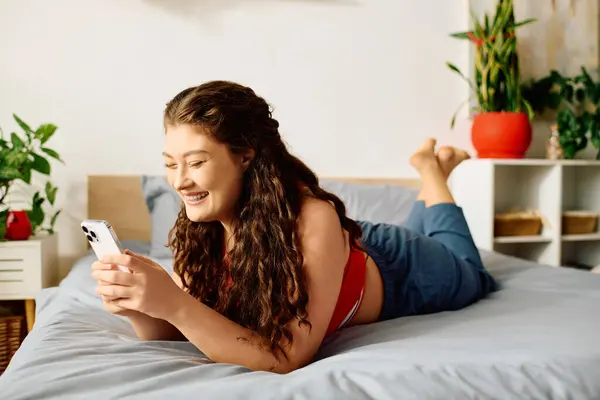 Eine fröhliche Plus-Size-Frau lümmelt bequem auf ihrem Bett, tief mit ihrem Telefon beschäftigt, während sie von Wohnkultur umgeben ist. — Stockfoto