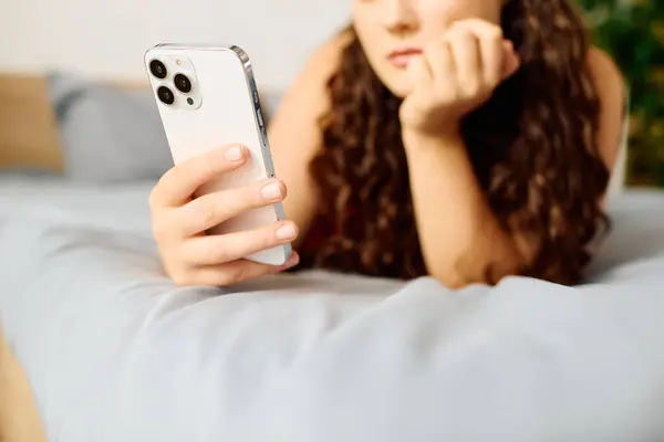 A young plus size woman with beautiful curly hair lounges comfortably on her bed, absorbed in her phone. — Stock Photo