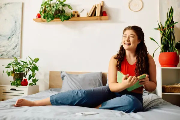 The young woman smiles joyfully, absorbed in her book while comfortably lounging on her bed surrounded by greenery. — Stock Photo