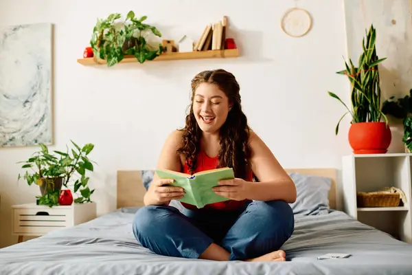 Una giovane donna con i capelli ricci sorride mentre si siede sul suo letto, godendo di un buon libro in una stanza verde, piena di piante. — Foto stock