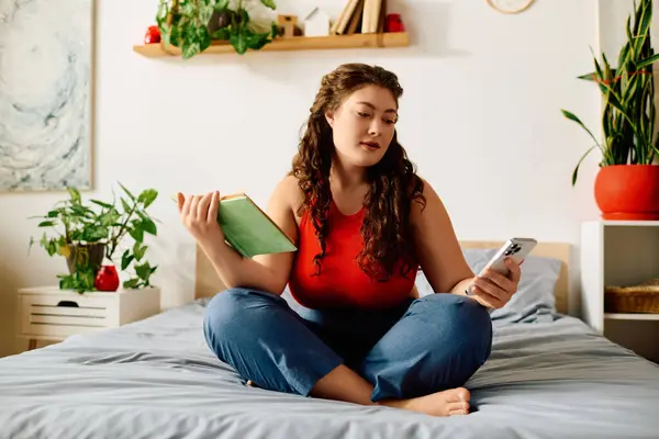 Relaxing at home, a young plus size woman with curly hair reads a book while checking her phone on her bedroom bed. — Stock Photo