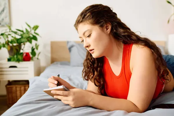 Eine junge Plus-Size-Frau mit schönen lockigen Haaren entspannt sich auf ihrem Bett und notiert ihre Gedanken in einem Notizbuch zu Hause.. — Stockfoto