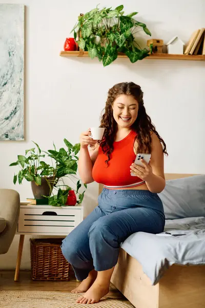 A plus size woman with curly hair relaxes at home, sipping tea and smiling while using her phone, surrounded by plants. — Stock Photo