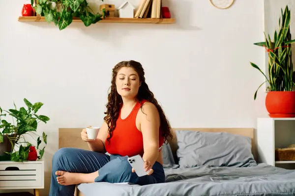 Relaxing in a bright and inviting room, a young plus size woman sips coffee while using her phone on the bed. — Stock Photo