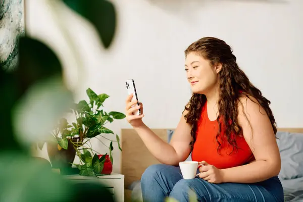 A beautiful plus-size woman relaxes at home, sipping tea while engrossed in her phone, surrounded by plants. — Stock Photo
