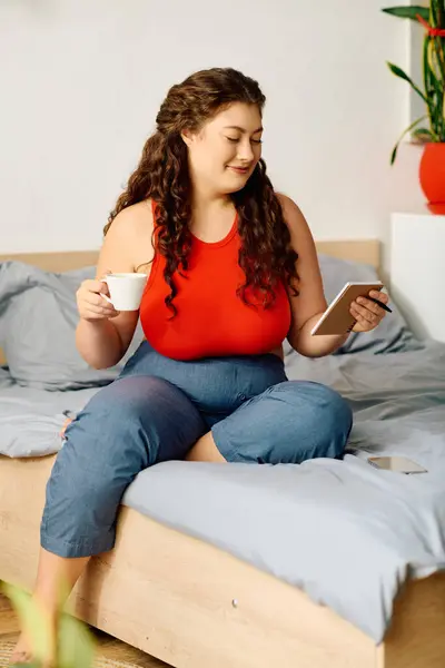 A beautiful plus-size woman with curly hair sits comfortably on her bed, sipping coffee and checking her phone, enjoying a cozy morning. — Stock Photo