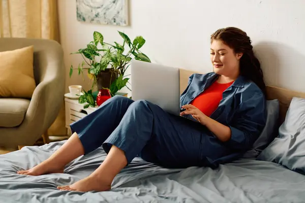 Una hermosa mujer de tamaño grande se relaja en su cama en casa, interactuando con su computadora portátil en un entorno cálido y acogedor. — Stock Photo