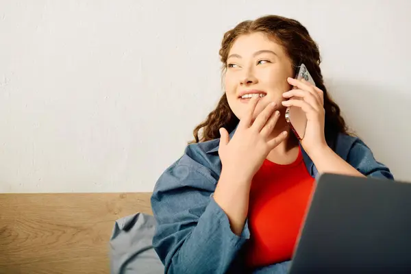 A cheerful young woman relaxes at home, chatting on the phone while comfortably using her laptop. — Stock Photo