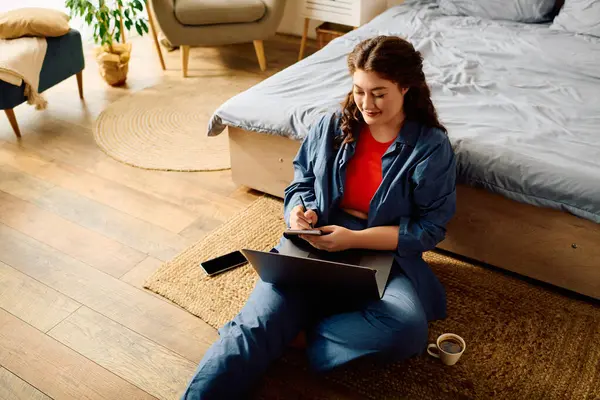 Веселая женщина plus size с кудрявыми волосами удобно сидит на полу, работая на ноутбуке и наслаждаясь уютной атмосферой дома. — Stock Photo