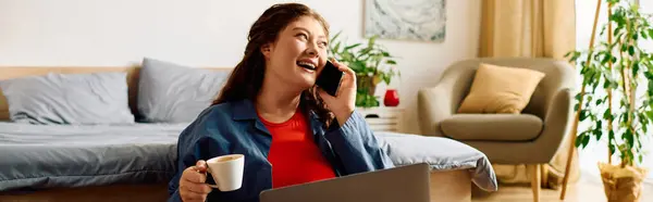 Una giovane donna plus-size con i capelli ricci chiacchiera gioiosamente al telefono, tenendo una tazza di caffè nel suo accogliente spazio abitativo. — Foto stock