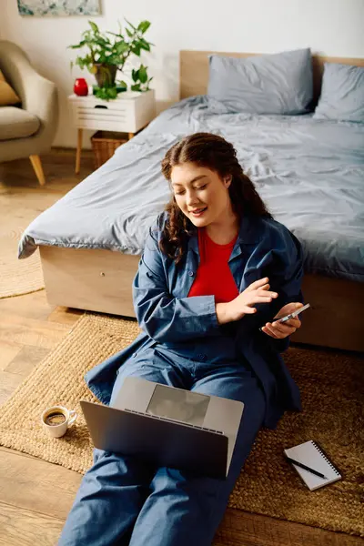 Una giovane donna plus size con i capelli ricci si siede sul pavimento, utilizzando il suo computer portatile e telefono mentre circondato da un ambiente accogliente casa. — Foto stock