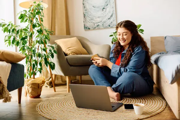 A young plus-size woman smiles while sitting on a cozy rug, engaging with her laptop in a bright, welcoming living space. — Stock Photo