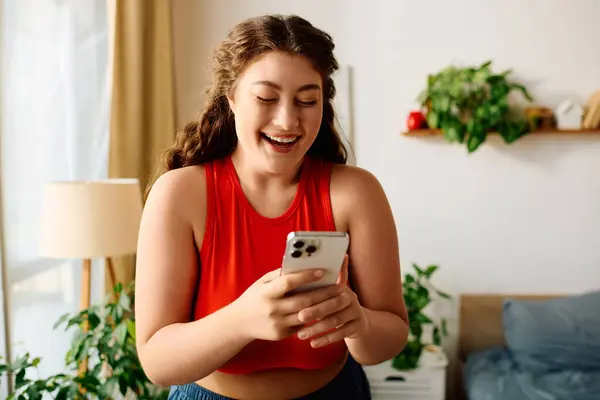 A beautiful plus size woman with curly hair smiles broadly while browsing her smartphone in a cozy home setting. — Stock Photo
