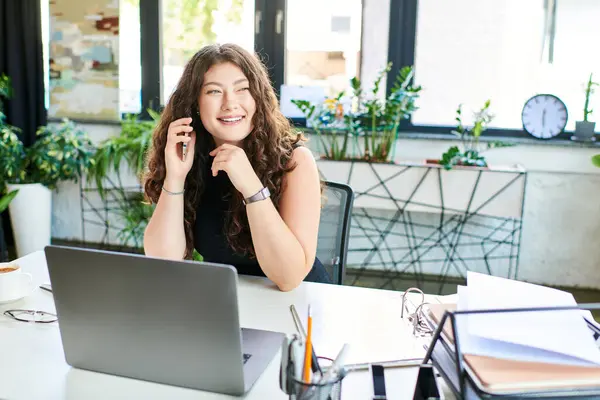A cheerful young woman with long curly hair engages in a phone call while working at her stylish office desk. — Stock Photo