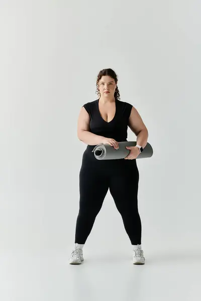 A confident plus size woman stands tall with a yoga mat, ready for practice in bright space. — Stock Photo