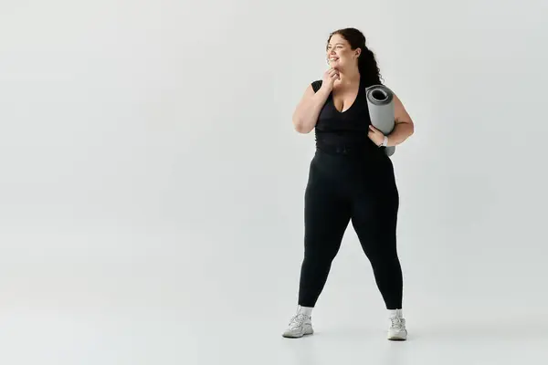 A confident plus size woman stands with a yoga mat, smiling in a bright, minimalistic room. — Stock Photo