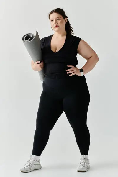 Una elegante mujer de talla grande se encuentra de pie, sosteniendo su esterilla de yoga, irradiando confianza y fuerza. - foto de stock