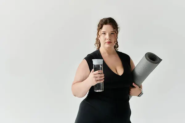 Una donna sicura di sé tiene una bottiglia d'acqua e un tappetino da yoga, pronti per il suo allenamento in studio. — Foto stock