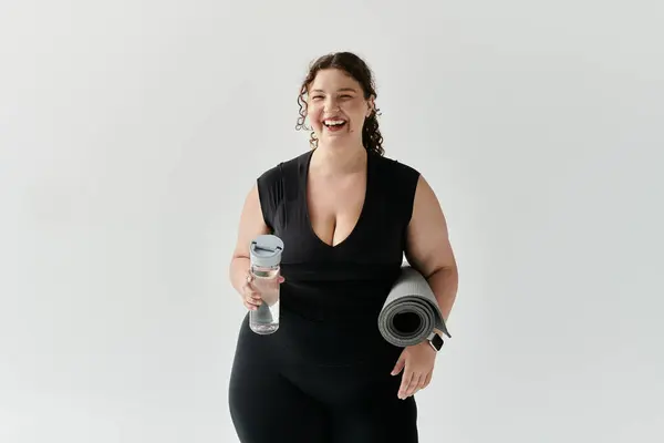 A joyful plus size woman in activewear holds a water bottle and yoga mat, radiating happiness. — Stock Photo