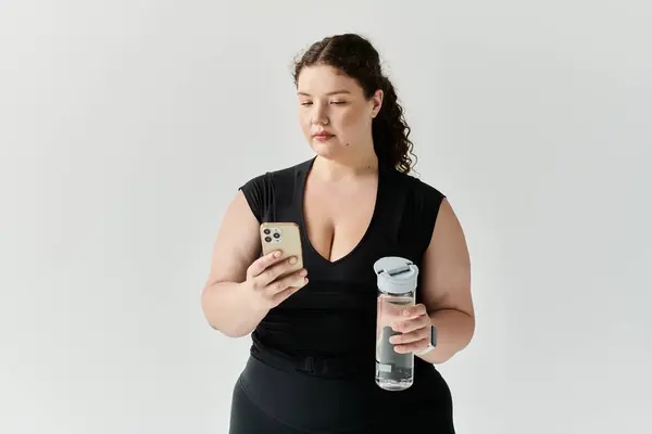 A beautiful plus size woman stands elegantly, focusing on her smartphone with a water bottle nearby. — Stock Photo